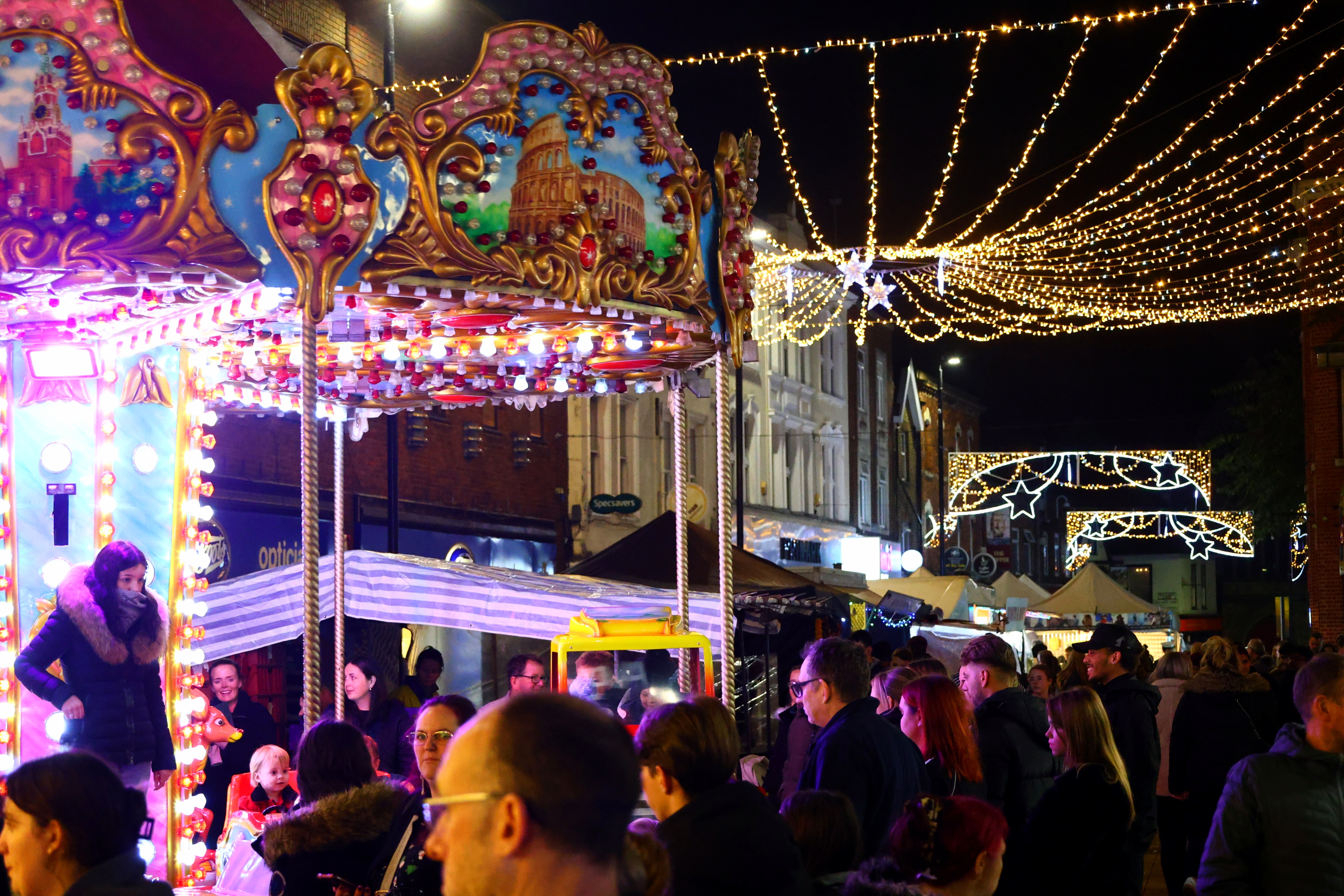 A high street at night with a brightly coloured carousel covered in lights, and christmas lights between shops.