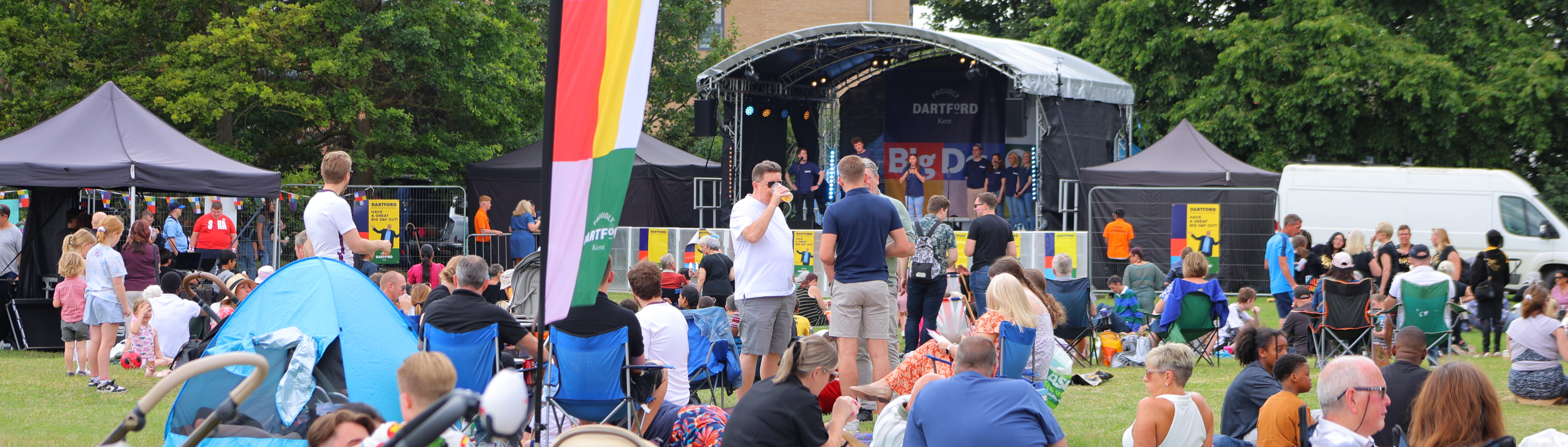 A stage with performers with people sat on the grass infront in chairs and on picnic blankets